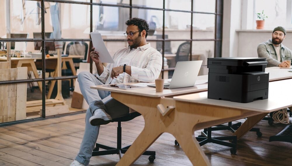 Concentrated young man in eyeglasses reading documents on paper while sitting at table with laptop and tablet near colleagues with computer during working in office