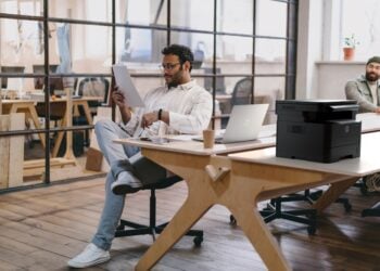 Concentrated young man in eyeglasses reading documents on paper while sitting at table with laptop and tablet near colleagues with computer during working in office